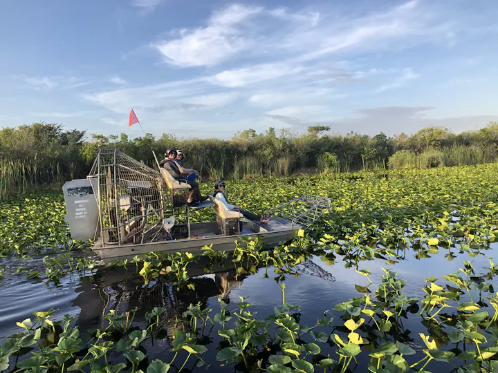 Picture of researchers in an airboat on shallow water with lily pads and trees in the background